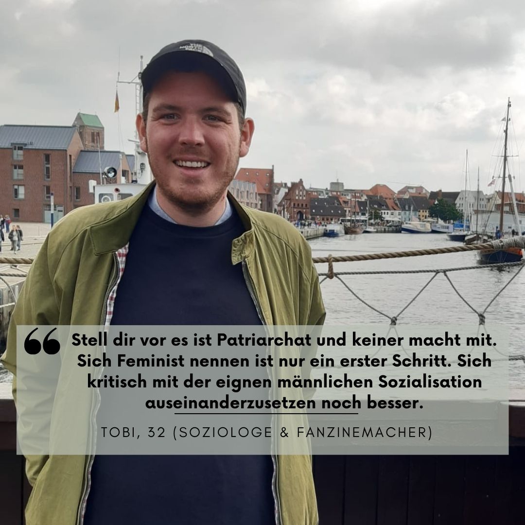 Junger mann, 32, mit Kappe und grünem Hemd steht vor einem Hafen auf einer Brücke und lächelt in die Kamera. Seine Hände hat er in den hinteren Hosentaschen stecken.